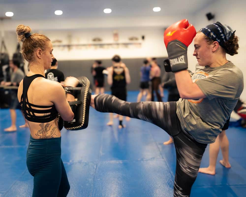 Two women training, one holding a pad and the other kicking it with red gloves on