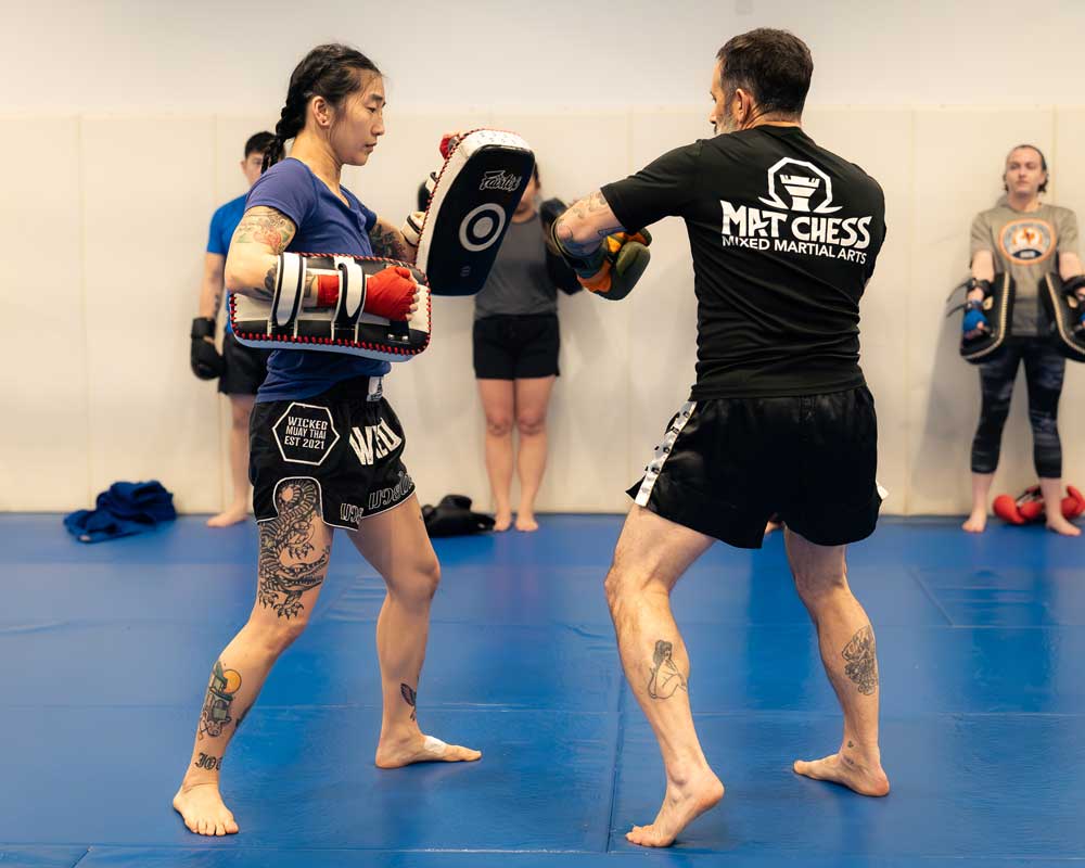 Two people training with focus mitts, one in a blue shirt and the other in black, while others watch