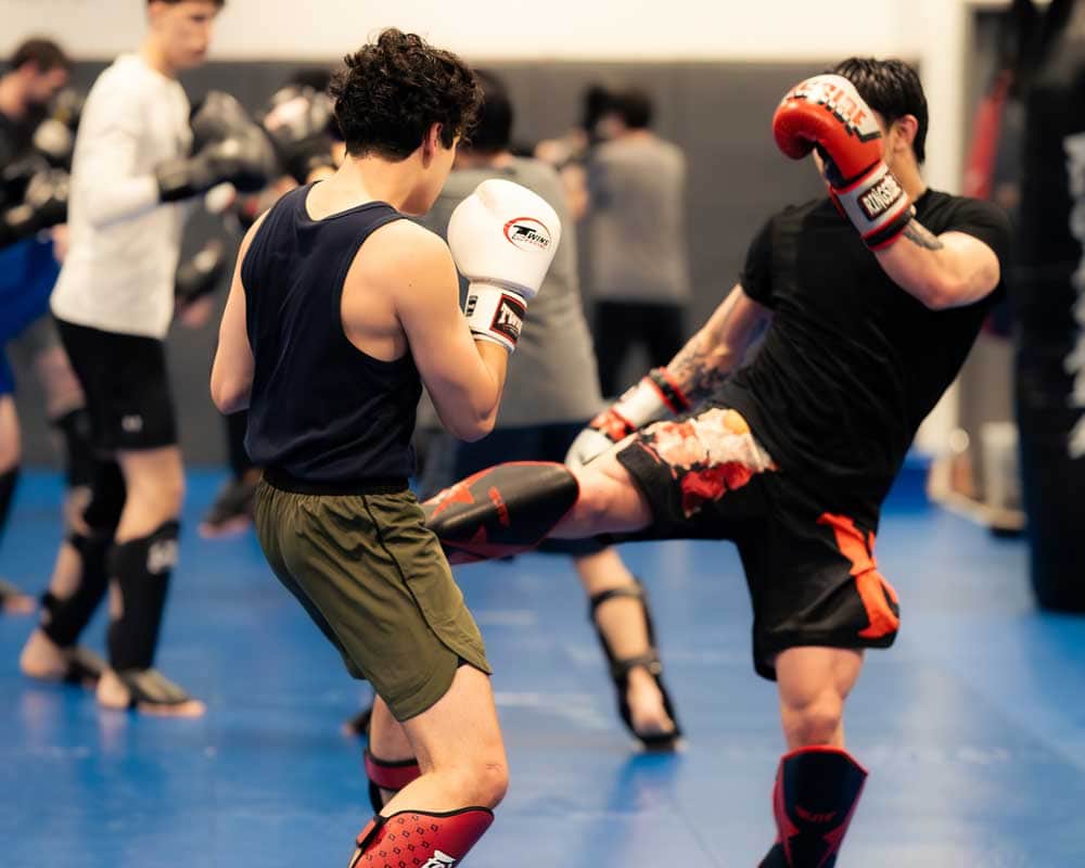 Two guys sparring in a gym, one throwing a kick and the other ready to block