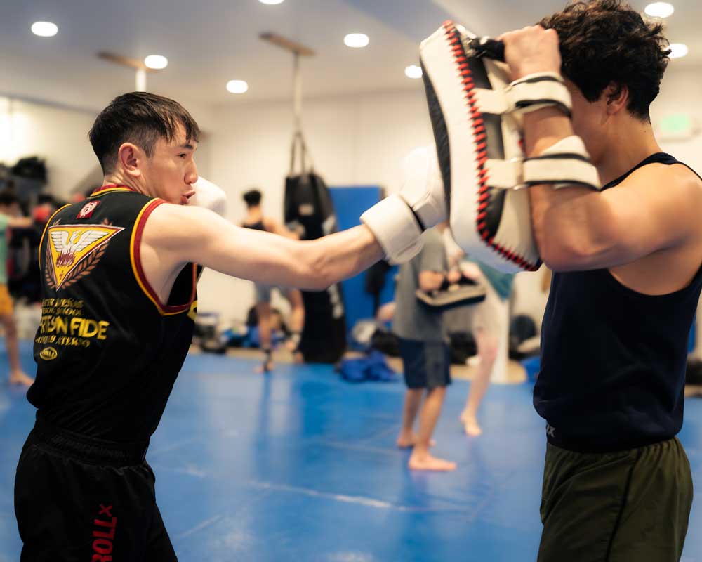 Guy in a black tank top throwing a punch at a focus mitt held by another guy in a gym