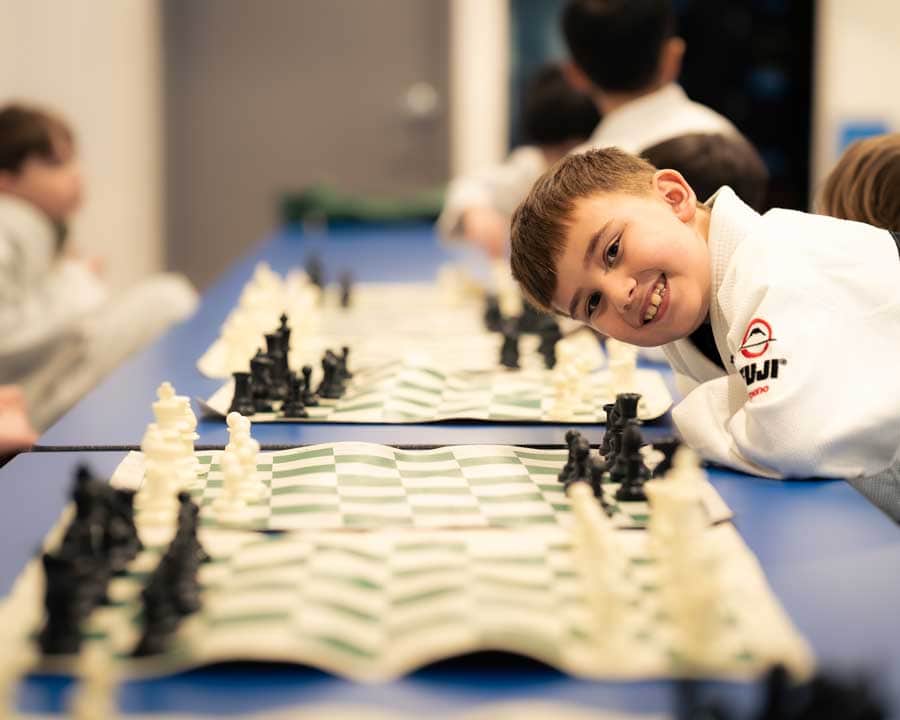 Kid in a white gi smiling while playing chess with other kids in the background