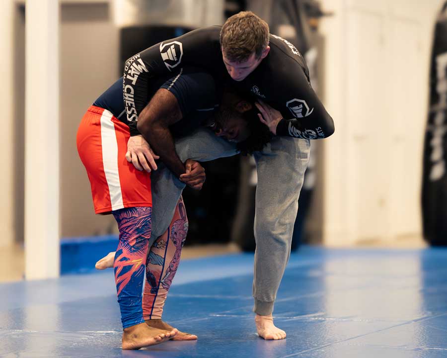 Two guys grappling on a gym mat, one bending down while the other holds him from behind