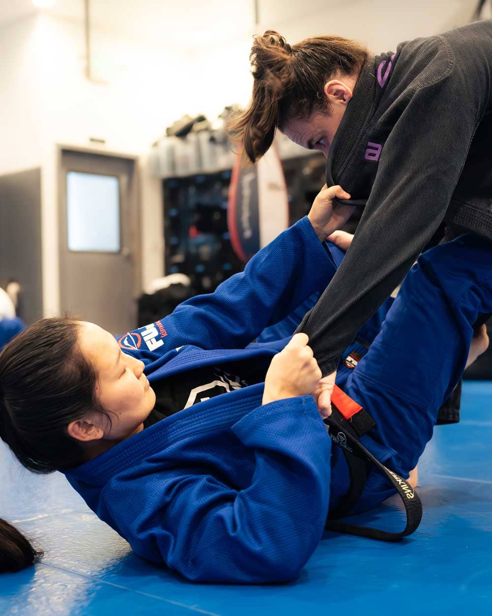 Two women sparring in jiu-jitsu, one on her back gripping the other's gi