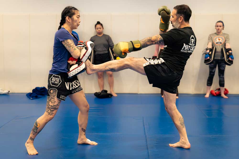 Man throwing a high kick at a woman holding pads during a martial arts practice