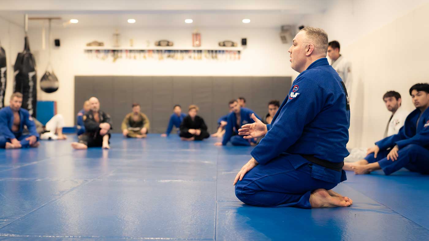 Guy in a blue gi kneels on a blue mat while others sit around listening